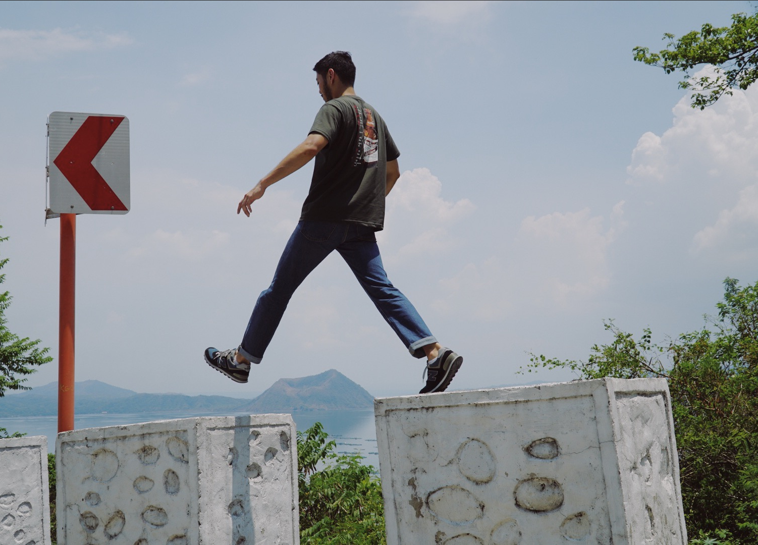 Image of paulo in mid-step as he appears to hop across concrete barriers. In the background, Taal Volcano can be seen in the distance across a body of water, under a clear blue sky scattered with clouds.